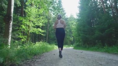 Genç bir bayan koşucu yaz ormanında antrenman yapıyor. Fitness kızı dışarıda koşuyor. Eğitim ve sağlıklı bir yaşam tarzı. Yavaş çekim.