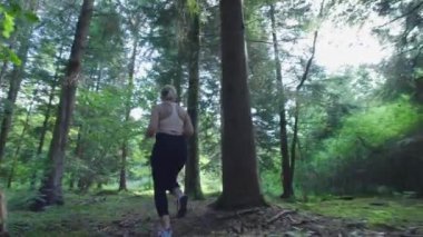 Genç bir bayan koşucu yaz ormanında antrenman yapıyor. Fitness kızı dışarıda koşuyor. Eğitim ve sağlıklı bir yaşam tarzı. Yavaş çekim.