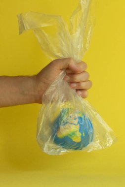 Bir adamın eli, dünyayı sarı bir zemin üzerinde plastik şeffaf bir torba içinde tutuyor, Dünya Günü için ekoloji konulu soyutlama, çevresel sorunlar, gezegenin plastik poşetlerle kirlenmesi