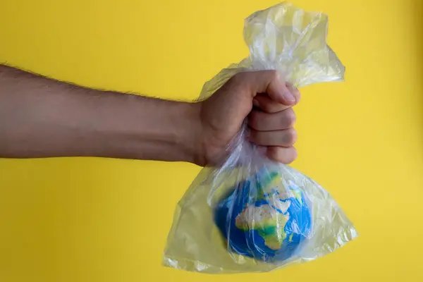 Bir adamın eli, dünyayı sarı bir zemin üzerinde plastik şeffaf bir torba içinde tutuyor, Dünya Günü için ekoloji konulu soyutlama, çevresel sorunlar, gezegenin plastik poşetlerle kirlenmesi