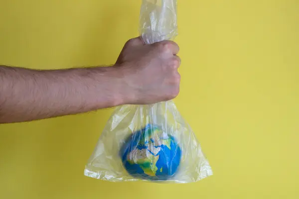 a man\'s hand holds the globe in a plastic transparent bag on a yellow background, abstraction on the theme of ecology for Earth Day, environmental problems, pollution of the planet with plastic bags