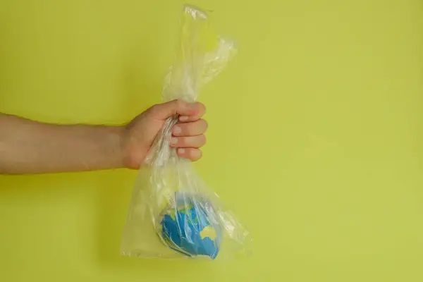 a man\'s hand holds the globe in a plastic transparent bag on a yellow background, abstraction on the theme of ecology for Earth Day, environmental problems, pollution of the planet with plastic bags