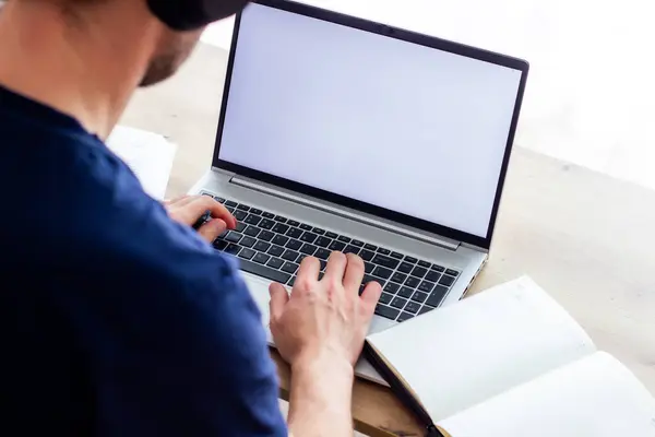 Modern kablosuz kulaklıklı bir adam gümüş bir dizüstü bilgisayarla yazı yazıyor, yakınlarda bir defter, boş beyaz ekranın önünde, küçük bir masada, açık renkli bir resim, online ders, toplantı, kafe, ofis.