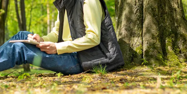 stock image a man in jeans and a vest draws with a simple pencil while sitting on the ground in the forest