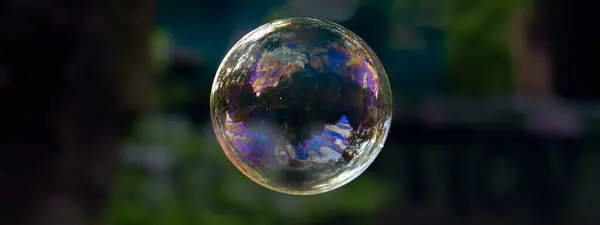 Mükemmel yuvarlak bir sabun köpüğü, mavi bir renk, güneş yansıması ve karanlık bir arkaplanda ışık parıltısı