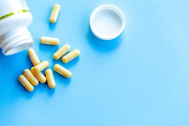 Bir kavanozun yanında bir avuç dolusu beyaz tıbbi kapsül ve parlak mavi bir arka plan.