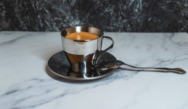 Zengin kahveyle dolu metalik bir fincan parlak bir kaşığın yanında parlak bir fincan üzerinde oturur. Mermer masa pürüzsüz bir arkaplan sağlar, ince görünümü geliştirir.