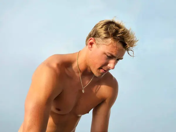 Shirtless millennial man working summer job at the beach against light blue sky with emotion. 