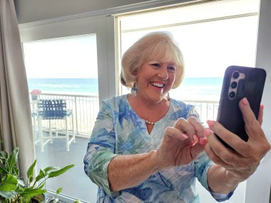 Happy senior grandma video chatting her grandkids on beach vacation in Florida.  clipart