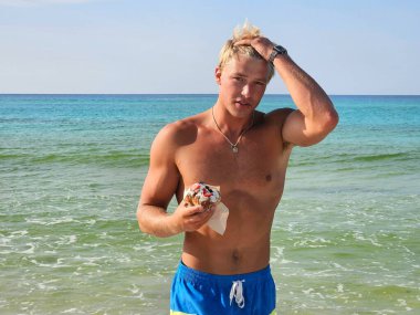 Genç adam Millennial Florida sahilinde kahvaltı niyetine çörek yerken sarışın saçlarını tarıyor..