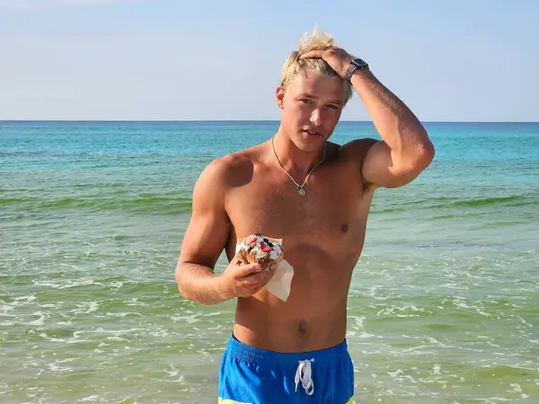 Genç adam Millennial Florida sahilinde kahvaltı niyetine çörek yerken sarışın saçlarını tarıyor..