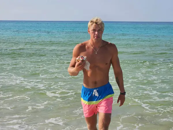 stock image Young millennial handsome dude in swim suit leaving ocean on beach vacation eating a Doughnut for breakfast. 