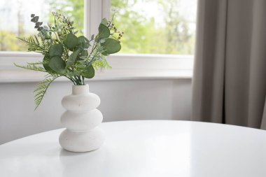 Beyaz masa üzerinde suni yeşillik ve arkasında pencere olan beyaz vazonun arka planı