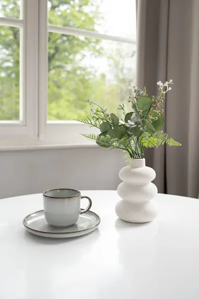 White vase with artificial greenery and empty cup on white table and window at the back
