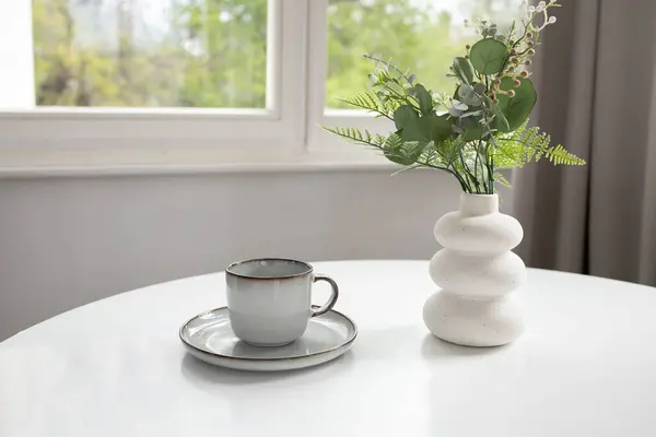 White vase with artificial greenery and empty cup on white table and window at the back
