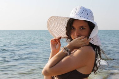 Genç bronzlaşmış kadın deniz kenarında mayo ve şapkayla poz veriyor.