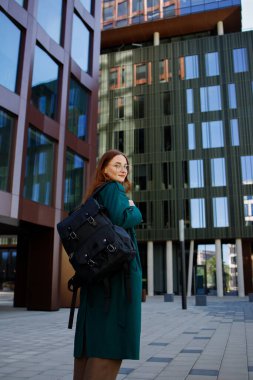 Kızıl saçlı kız öğrenci mi yoksa gözlüklü işçi mi? Sırt çantalı ve endüstriyel binalarla çevrili.