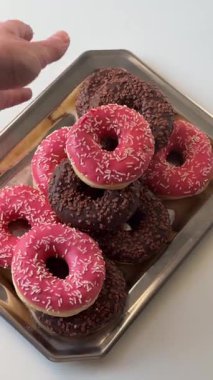 Kadın eli, metal tepsiden pembe bir çörek alıyor. Bir sürü pembe ve kahverengi çörek.