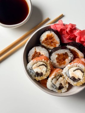 Suşi, pembe zencefil, soya sosu ve beyaz masada tahta çubuklarla dolu bir kasede yuvarlanıyor.