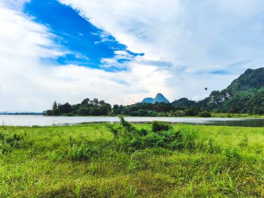 A beautiful Scenic view of the mountain with green grass and a calm lake with cloudy blue sky background at tasik timah tasoh, perlis, malaysia. clipart
