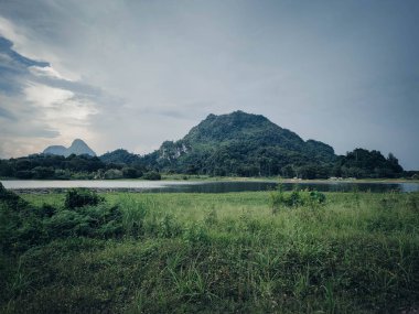 A dark and beautiful Scenic view of the mountain with green grass and a calm lake with cloudy blue sky background at tasik timah tasoh, perlis, malaysia. clipart