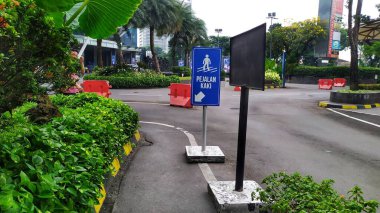 Alış veriş merkezinin girişinin yanında üzerinde beyaz yazı ve insan sembolü olan mavi bir yaya levhası. Yaya levhasının solunda küçük bir bahçe. Jakarta, Endonezya, 21 Mayıs 2024.