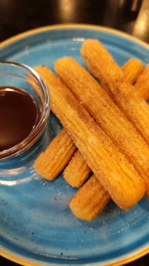 Tabakta çuroslar cezbedici, altın kahverengidir ve şekerle kaplıdır, yanında da sos vardır. Lezzetli çuro yığını mavimsi yeşil bir tabakta duruyor..