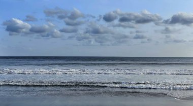 Tropikal bir kıyı şeridi boyunca, ufuk geniş mavi gökyüzüne açılırken küçük dalgalar yavaşça kumda süzülür, beyaz-gri bulutlar ılık esintide yavaşça sürüklenir.. 