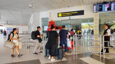 Uçak yolcuları 2 Temmuz 2025 'te I Gusti Ngurah Rai Uluslararası Havalimanı, Bali, Endonezya' ya giriş yapmak için iç kalkış bölgesine doğru yola çıkmaktadır. Girişte, erkek bir güvenlik görevlisi nöbet tutuyor, düzeni ve güvenliği sağlıyor.