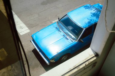 Captured from a window on a bright sunny day, this photo showcases a striking blue car illuminated by the warm rays of the sun, adding a vibrant touch to the scene clipart