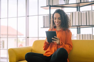 Latin kadın dijital tablet kullanıyor ve evdeki kanepede otururken gülümsüyor.