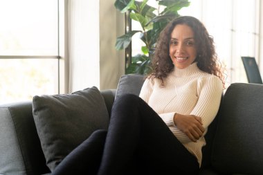 Latin kadın gülümsüyor ve evdeki kanepede oturuyor.