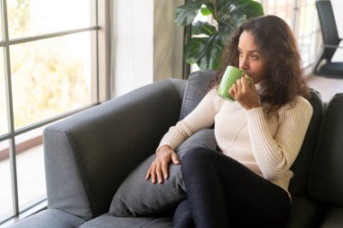 Latin kadın evdeki kanepede kahve içiyor.