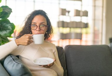 Latin bir kadın evdeki kanepede kahve içiyor. Eski moda efekt filtresi.