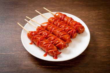 Beyaz tabakta ketçaplı kızarmış sosis şişi.