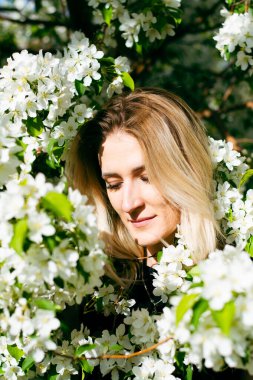 Parkta duran genç sarışın kadın İlkbaharda çiçek açmış elma ağacının yanında