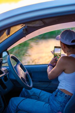 Kadın elinde gün batımında arabanın içinde fotoğraf çekiyor.
