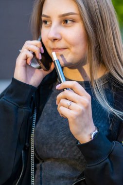 Sokakta e-sigara içen genç bir kadın telefonda konuşuyor. Bağımlılık konsepti. Yüksek kalite fotoğraf. Yüksek kalite fotoğraf
