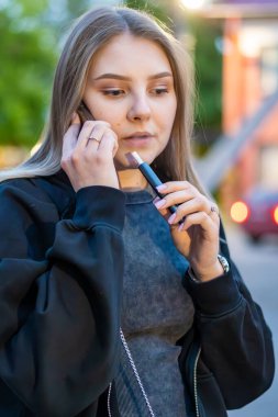 Sokakta e-sigara içen genç bir kadın telefonda konuşuyor. Bağımlılık konsepti. Yüksek kalite fotoğraf. Yüksek kalite fotoğraf
