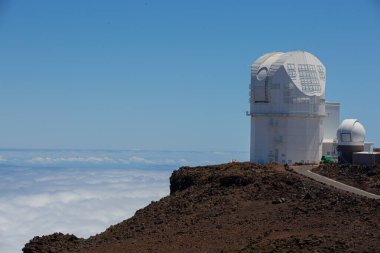 Haleakala kayalık zirvesinde, parlak mavi gökyüzünün altında sonsuz genişlikte bulutlarla çevrili, bilimsel gelişmeleri ve keşifleri ön plana çıkaran el değmemiş bir gözlemevi..