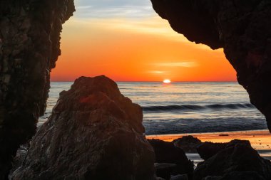 Malibu 'daki Matador Plajı' ndaki pürüzsüz kaya oluşumları tarafından çerçevelenmiş okyanus ufkuna batan parlak bir günbatımı. Alacakaranlık gökyüzünün ılık parıltısı altında yumuşak dalgalar ve dingin doğal güzellik..