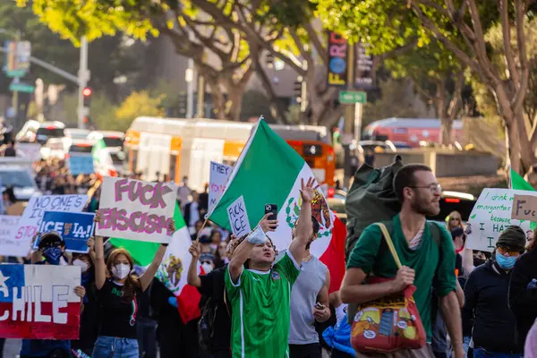 Muhtelif protestoculardan oluşan geniş bir kalabalık Los Angeles şehir merkezinde anti-ICE ve Trump karşıtı levhalarla Meksika ve Amerikan bayrakları sallayarak toplanıyor. Siyasi gösteri, göçmen hakları, sosyal adalet ve toplumsal dayanışmayı destekliyor. Fotoğraflı dur
