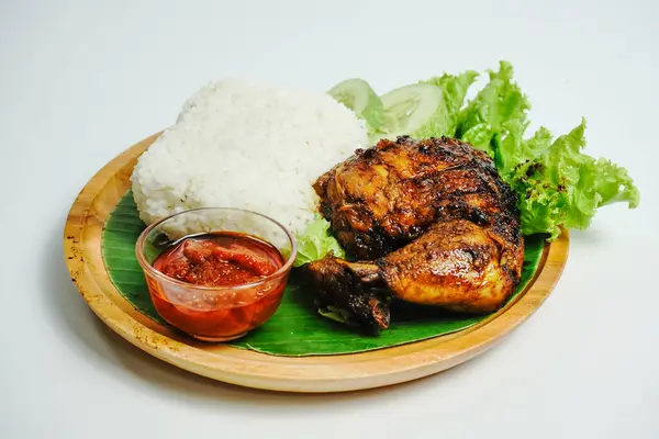 Grilled chicken served with rice, chili paste, slice of cucumber and lettuce leaves. Nasi ayam bakar lalapan, authentic recipe of Indonesian grilled chicken.
