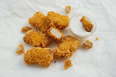 Fried crispy crumb chicken nuggets next to glass bowl of white sauce on white baking paper. Dipped nugget. Top view.                                clipart