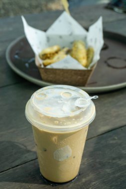 Plastik kapakla servis edilen buzlu kahveyle doldurulmuş şeffaf plastik bardak. Arka planda muzlu börek dolu bir sepet var..