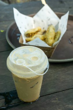 Plastik kapakla servis edilen buzlu kahveyle doldurulmuş şeffaf plastik bardak. Arka planda muzlu börek dolu bir sepet var..