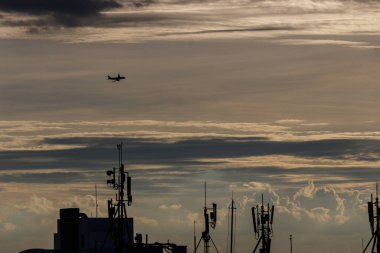 Günbatımında gökyüzünde uçan ticari bir uçakla şehir manzarası, binalar ve antenler arasında