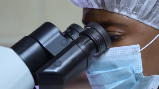 戴医疗面罩的非裔美国女人透过显微镜看 近景肖像 — 图库视频影像