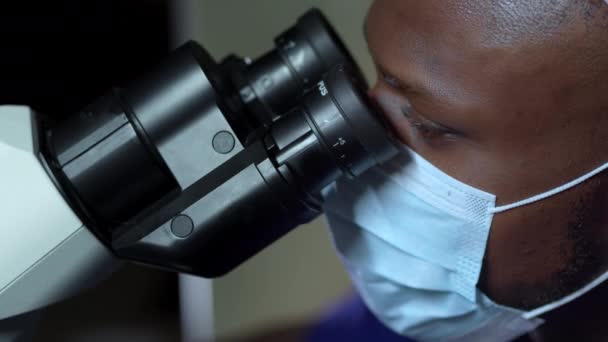 戴医疗面具的非裔美国人透过显微镜看 近景肖像 — 图库视频影像