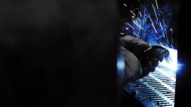 Fabrika işçisi metali kaynaklıyor. Adam kaynak yapıyor. Kaynak makinesi kullanarak argon veya elektrot ile kaynak yapmak. Metal yapılar üreten bir endüstriyel girişim. Kıvılcımlar ve parıltılar uçuşuyor. 